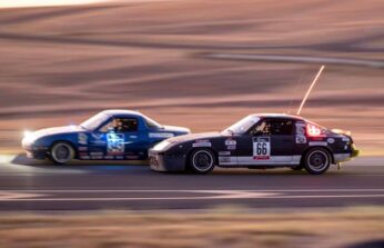 Gray Cloud Racing black #66 Mazda RX-7 battling side by side with a blue Mazda Miata during an endurance race at sunset